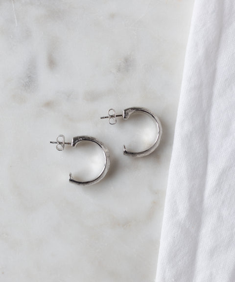 A pair of WALD Berlin SAMPLE SALE Pop Classy Hoop Silver earrings with stud backs are displayed on a white marble surface beside a white cloth.