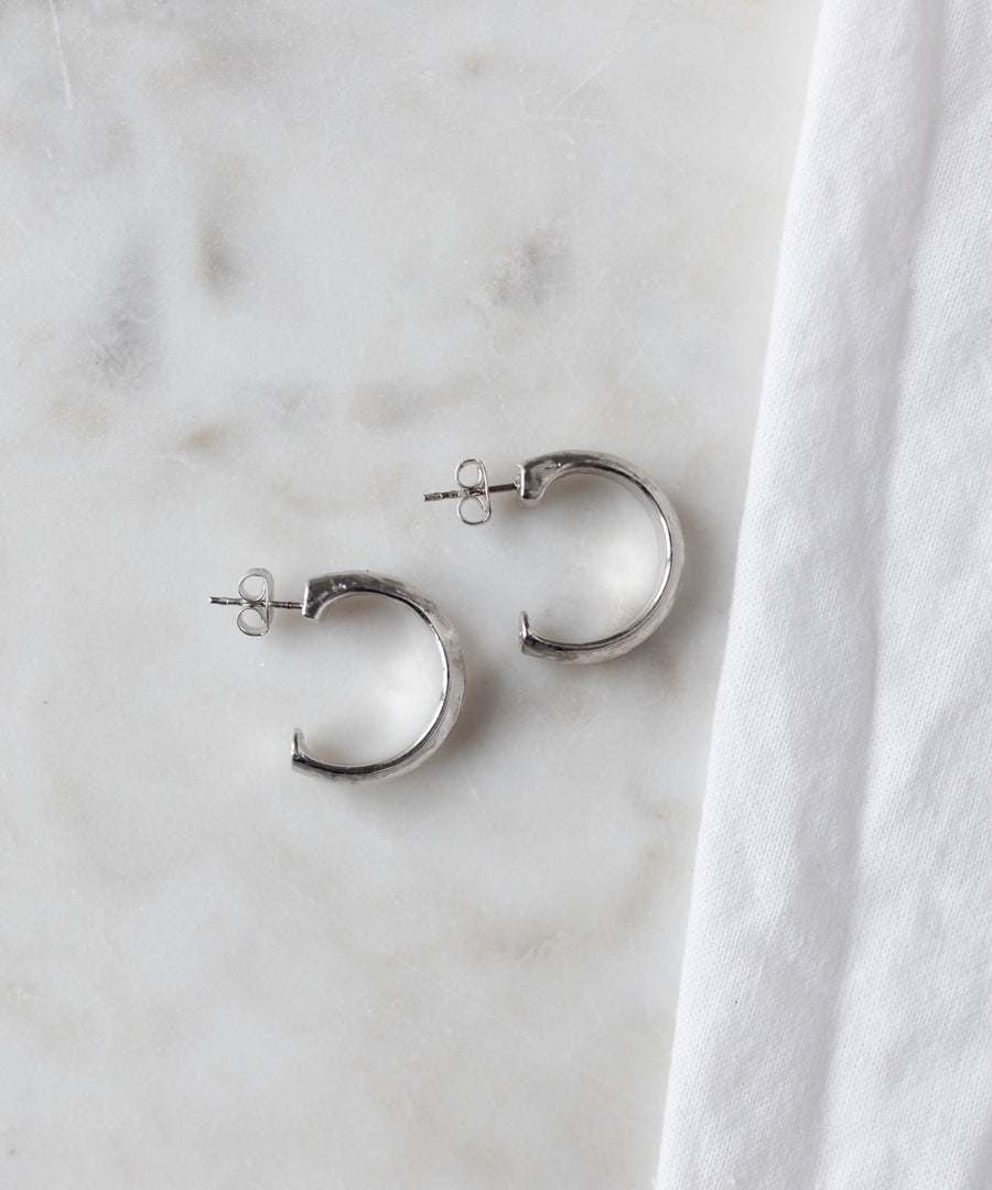 A pair of WALD Berlin SAMPLE SALE Pop Classy Hoop Silver earrings with stud backs are displayed on a white marble surface beside a white cloth.