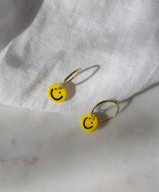 Two gold-plated hoop earrings from WALD Berlin, called Smilie Yellow Earrings, each feature a yellow smiley face charm and rest on a white textured fabric surface. Expertly crafted in Germany, these earrings bring a cheerful accent to any outfit.