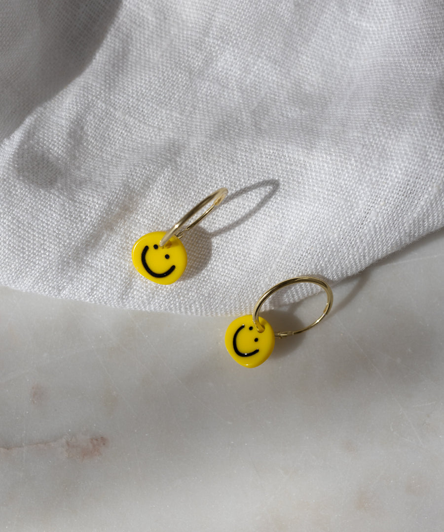 Two gold-plated hoop earrings from WALD Berlin, called Smilie Yellow Earrings, each feature a yellow smiley face charm and rest on a white textured fabric surface. Expertly crafted in Germany, these earrings bring a cheerful accent to any outfit.