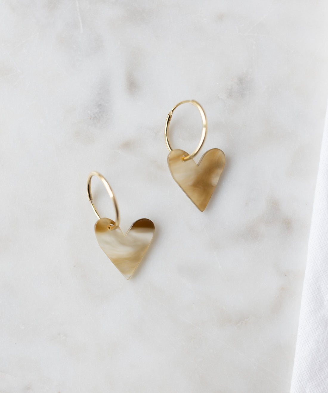 The WALD Berlin Exclusive Heart Black Earrings feature recycled gold hoops with marbled black heart-shaped pendants, elegantly displayed on a light marble surface.