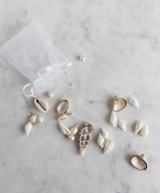 A sheer white drawstring bag from WALD Berlin’s Candyman Shell Kit spills handpicked seashell and pearl pendants—each a unique jewel piece—onto a marble surface.
