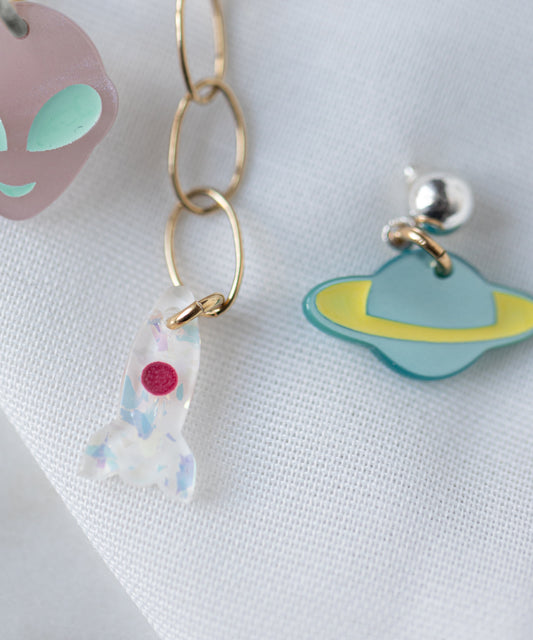 The WALD Berlin SAMPLE SALE From Another Mother Galactic Earring Set features playful bio polyester-accented acrylic charms—a purple alien face, white rocket, and blue planet—styled as earrings on a white fabric background.