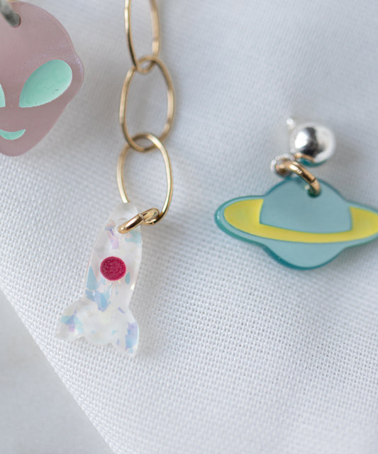 The WALD Berlin SAMPLE SALE From Another Mother Galactic Earring Set features playful bio polyester-accented acrylic charms—a purple alien face, white rocket, and blue planet—styled as earrings on a white fabric background.