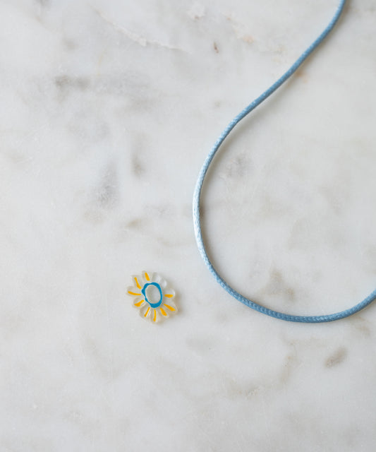 A Daisy Transparent Charm pendant with a turquoise center and a blue cord on a marble surface, ideal for DIY jewelry enthusiasts by WALD World.