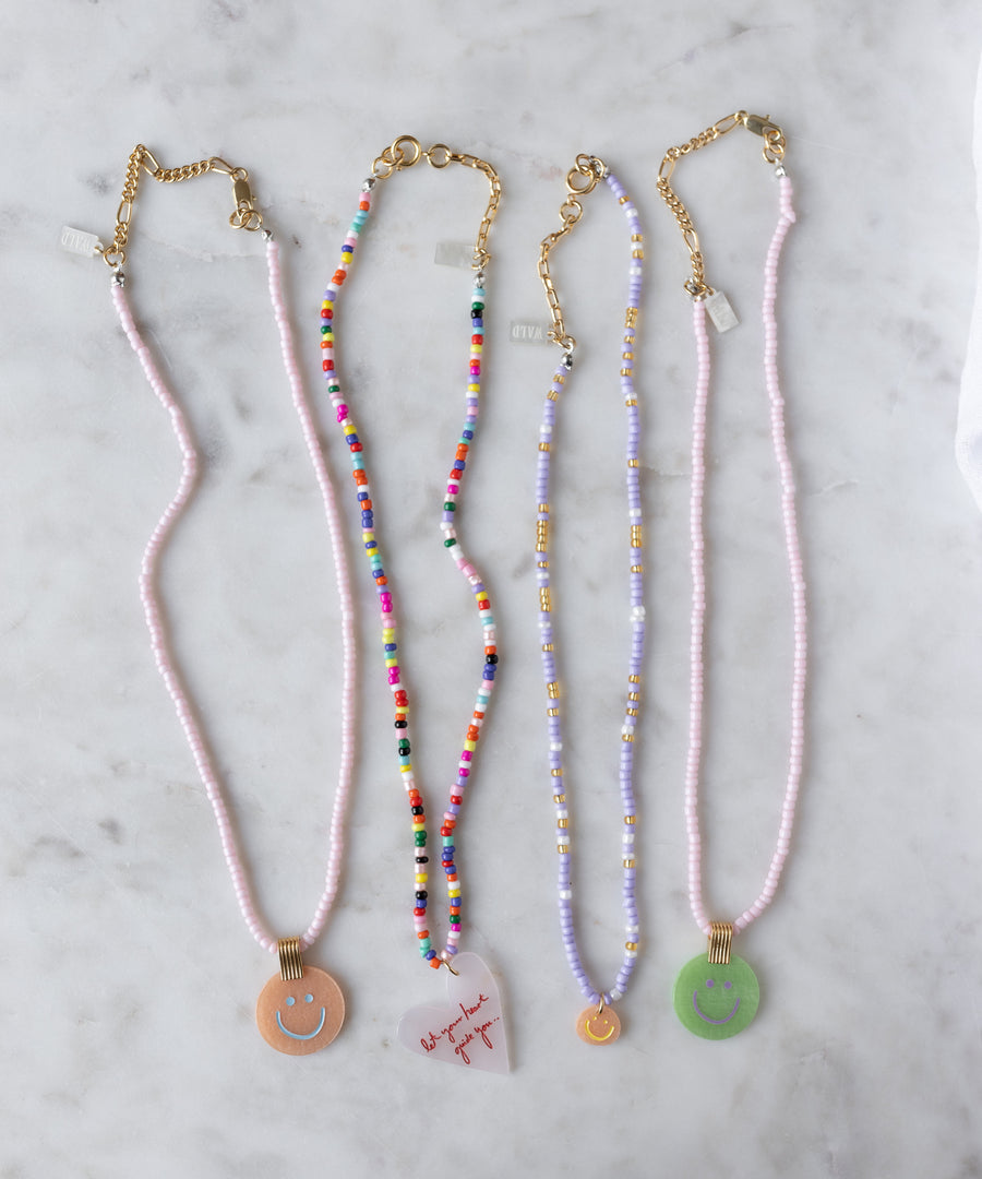 Four WALD Berlin necklaces with gold clasps rest on marble: three feature the Pop Smilie Medallion Peach pendant, while one is adorned with a heart and red text for a playful summer touch.