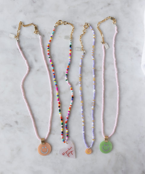 Four 18k gold plated beaded necklaces by WALD Berlin are displayed on marble; each features a unique summer pendant, such as the Pop Smilie Medallion Green and a heart charm.