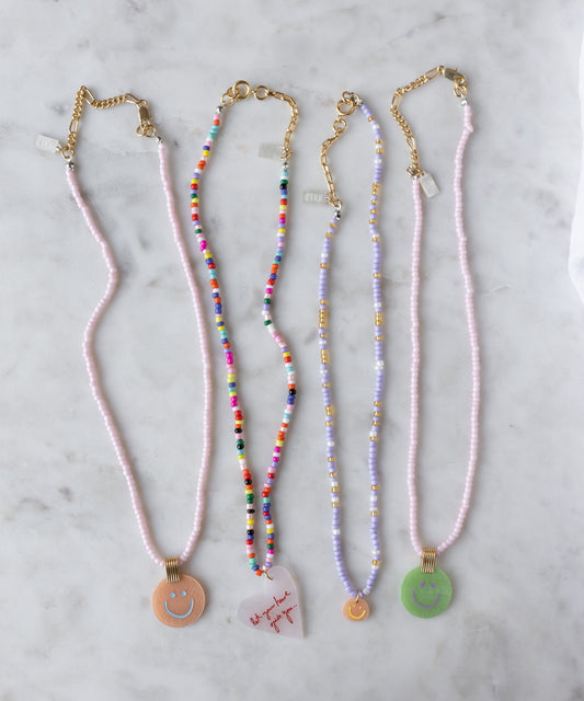 Four WALD Berlin Pop Smilie Mini beaded necklaces with 18k gold plated clasps are displayed on a marble surface, each featuring a unique pendant—including a smilie and a heart with text.