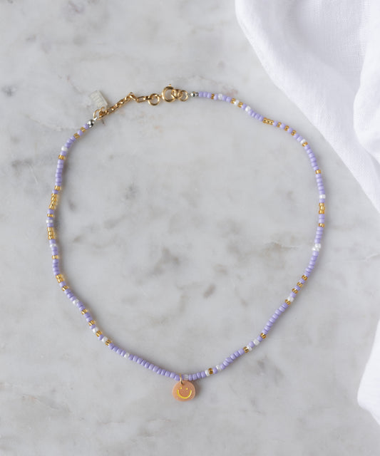 The Pop Smilie Mini necklace by WALD Berlin, made with light purple and 18k gold plated beads and a small round Smilie Medaillon pendant, is shown on marble beside a white cloth.