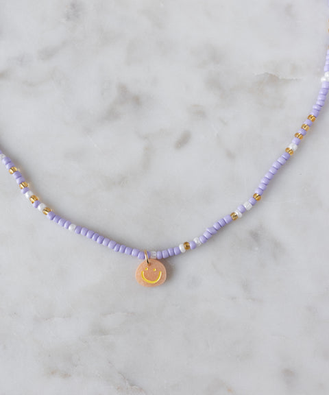 The WALD Berlin Pop Smilie Mini necklace features small purple and gold beads with an 18k gold plated Smilie Medaillon pendant, displayed on a light marble surface.