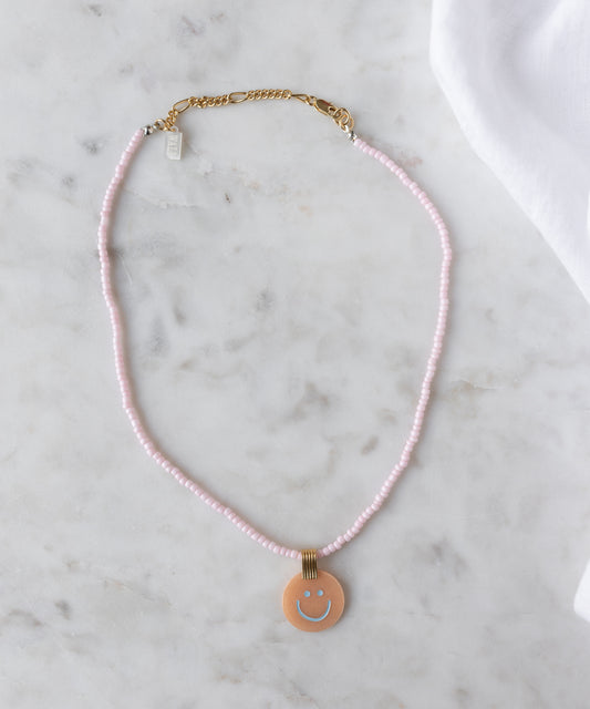 The Pop Smilie Medallion Peach by WALD Berlin is a pink beaded necklace with a gold clasp and round pendant featuring a blue smiley face—ideal for adding playful charm to classic styles or your summer look.