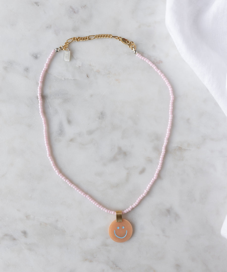 The Pop Smilie Medallion Peach by WALD Berlin is a pink beaded necklace with a gold clasp and round pendant featuring a blue smiley face—ideal for adding playful charm to classic styles or your summer look.