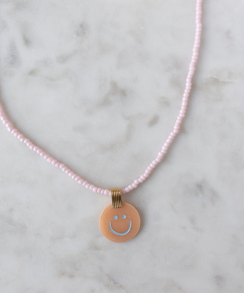 Add a cheerful pop to your style with the Pop Smilie Medallion Peach necklace from WALD Berlin—a pink beaded piece with a round peach pendant, blue smiley face, and playful vibes, perfect for brightening up your summer look.