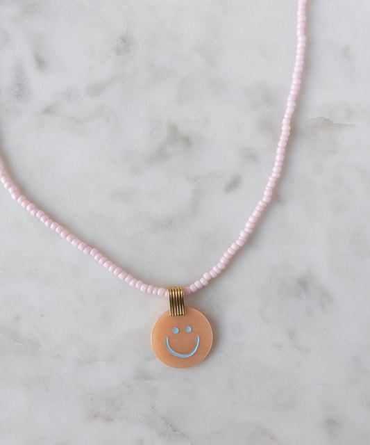 Add a cheerful pop to your style with the Pop Smilie Medallion Peach necklace from WALD Berlin—a pink beaded piece with a round peach pendant, blue smiley face, and playful vibes, perfect for brightening up your summer look.
