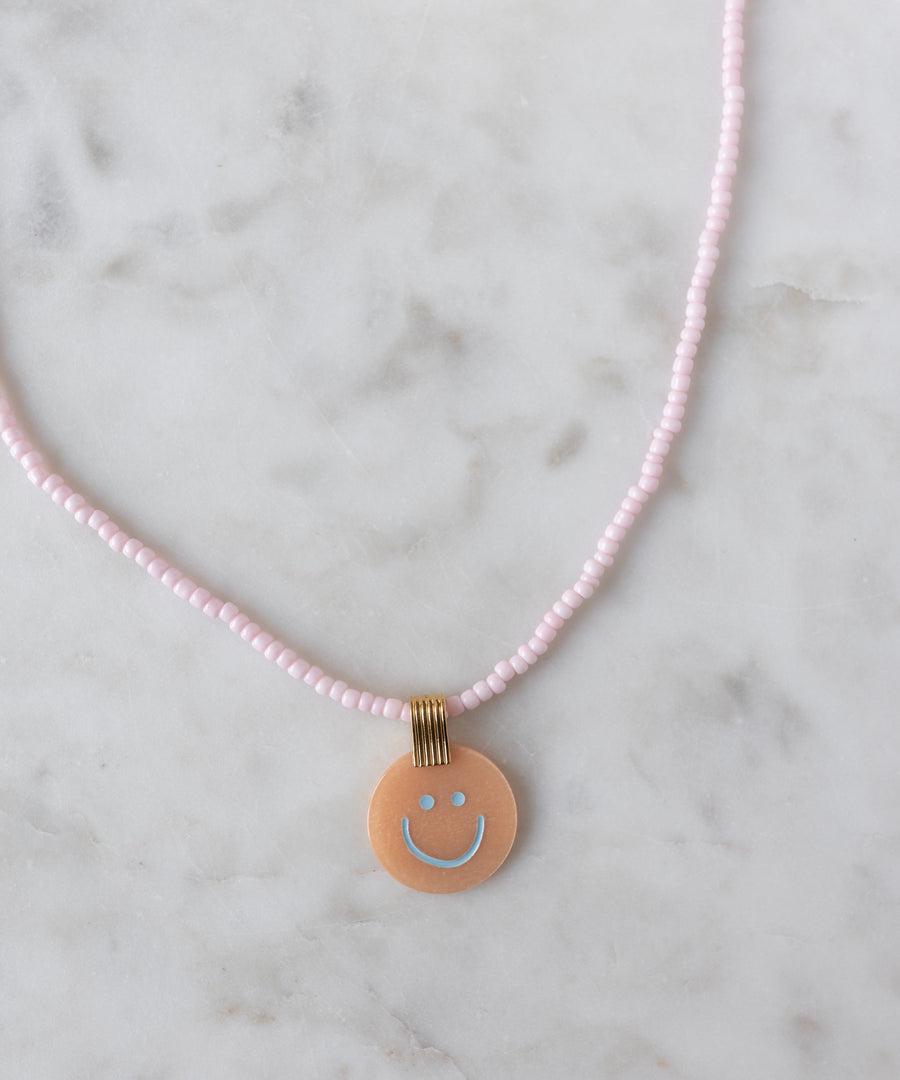 Add a cheerful pop to your style with the Pop Smilie Medallion Peach necklace from WALD Berlin—a pink beaded piece with a round peach pendant, blue smiley face, and playful vibes, perfect for brightening up your summer look.