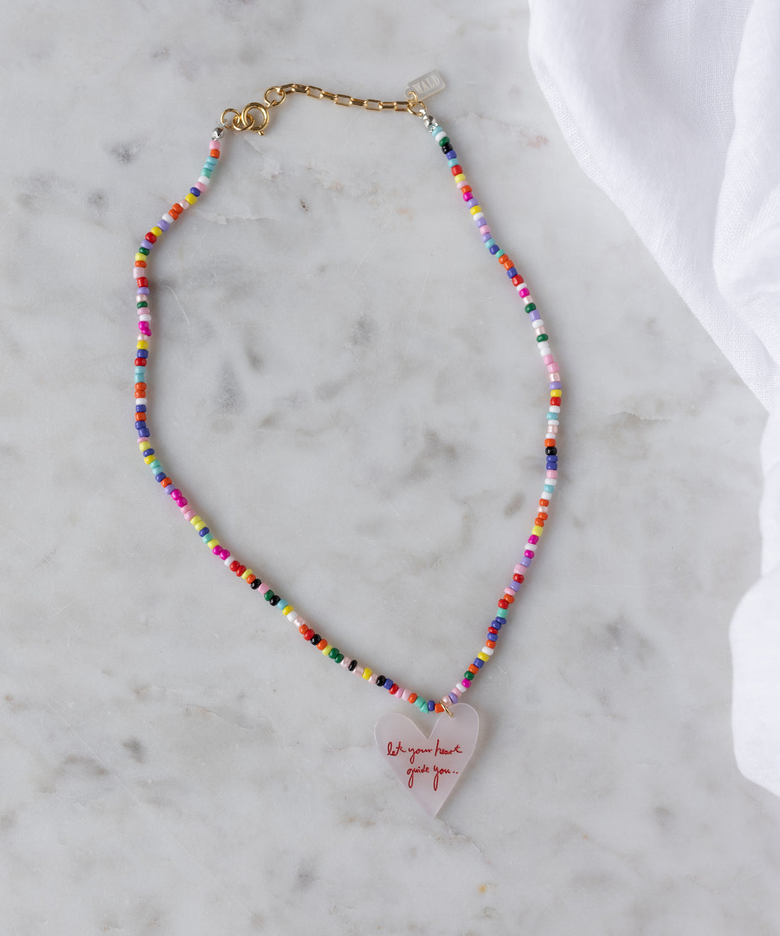 The WALD Berlin SAMPLE SALE Pop Let Your Heart Guide You necklace features 18k gold plated details, a heart-shaped pendant with red text, and a gold clasp, showcased on a marble surface.