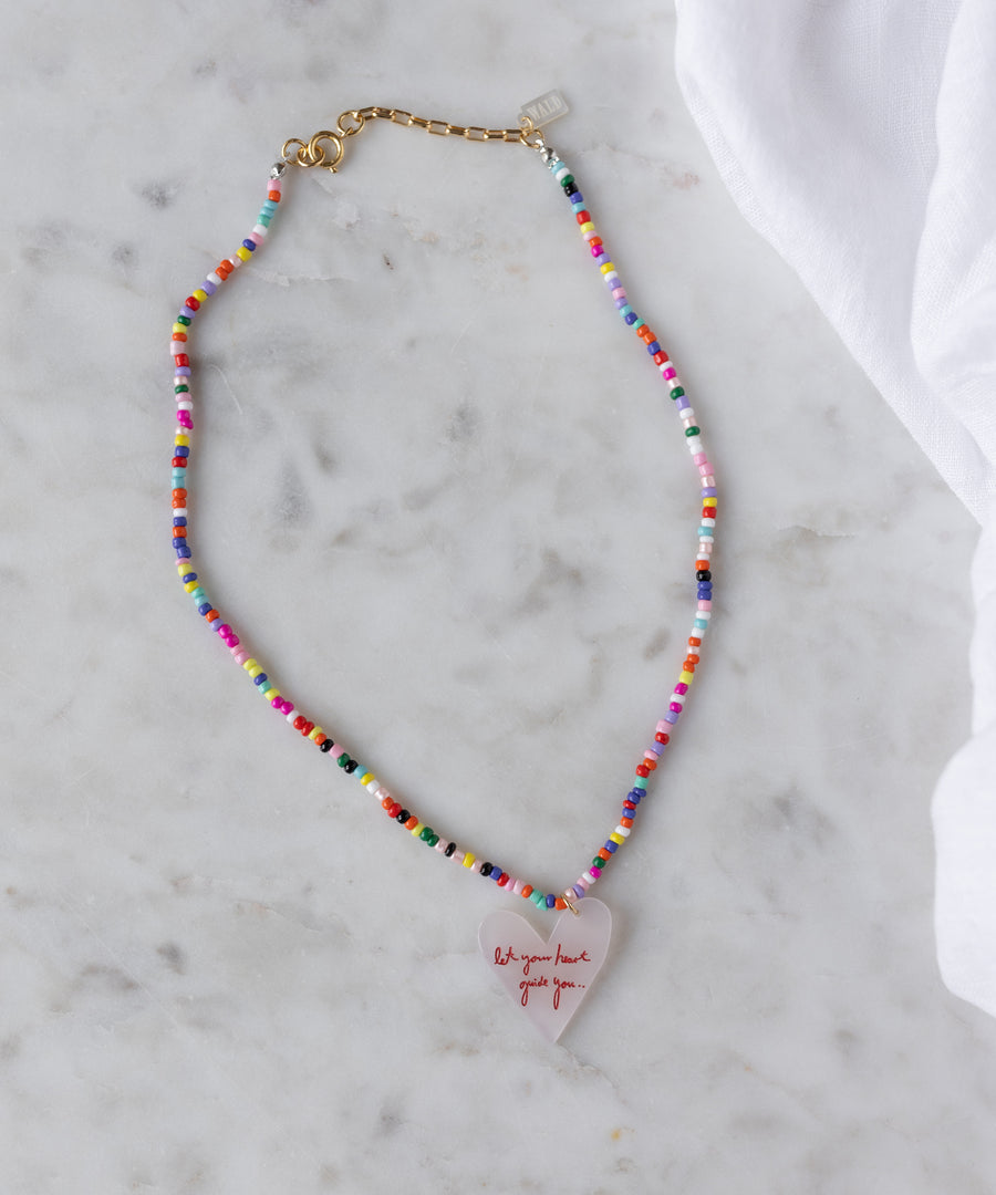 The WALD Berlin SAMPLE SALE Pop Let Your Heart Guide You necklace features 18k gold plated details, a heart-shaped pendant with red text, and a gold clasp, showcased on a marble surface.