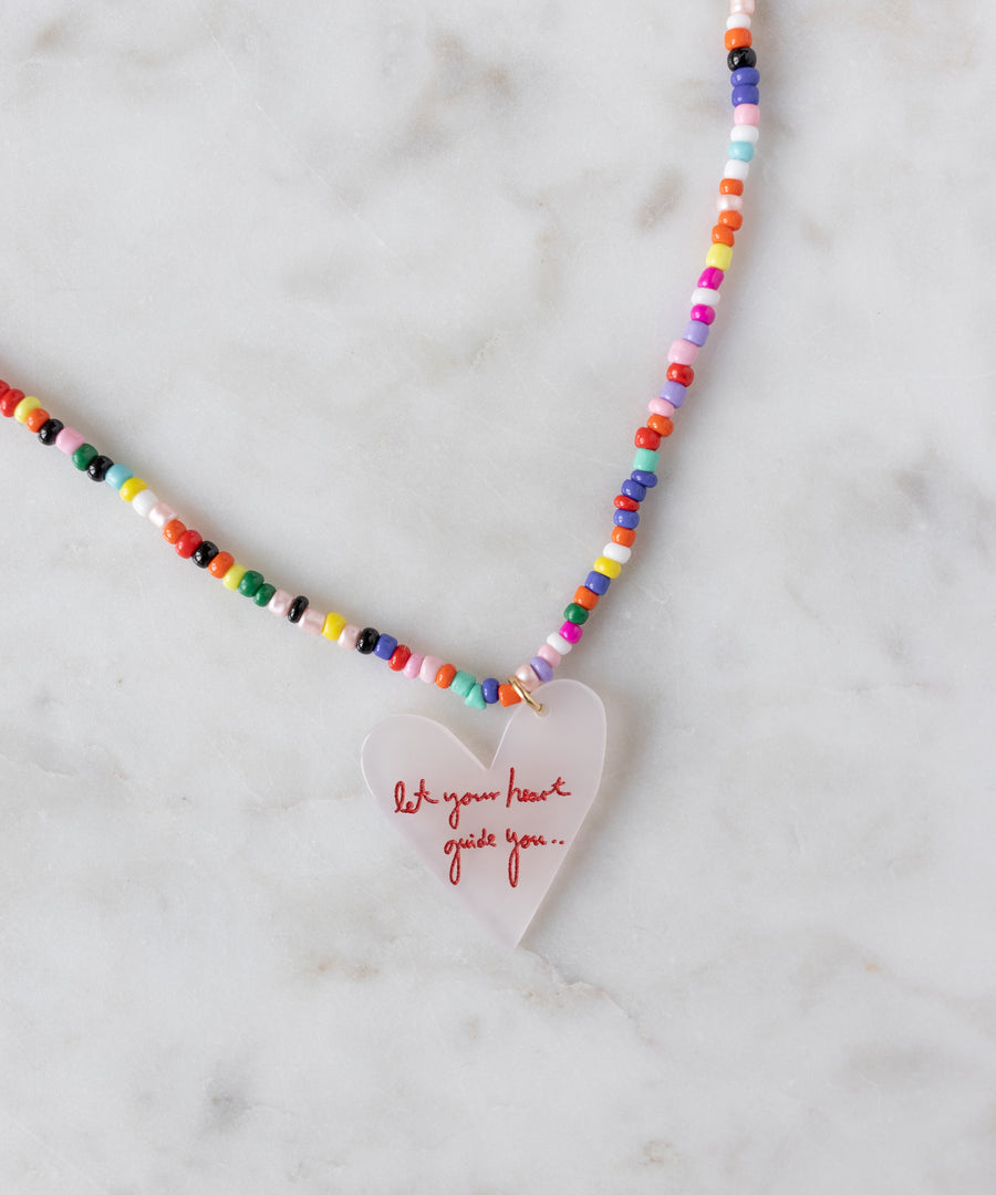 The WALD Berlin SAMPLE SALE Pop Let Your Heart Guide You necklace features colorful beads and an 18k gold plated heart pendant inscribed with "let your heart guide you..." in red, displayed on a marble surface.
