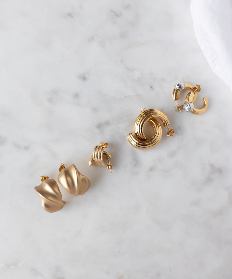 Four pairs of 18k gold plated earrings, including hoops and abstract shapes, from WALD Berlin's SAMPLE SALE Matt Gold Creoles collection are arranged on white marble—exclusive picks from our sample sale.