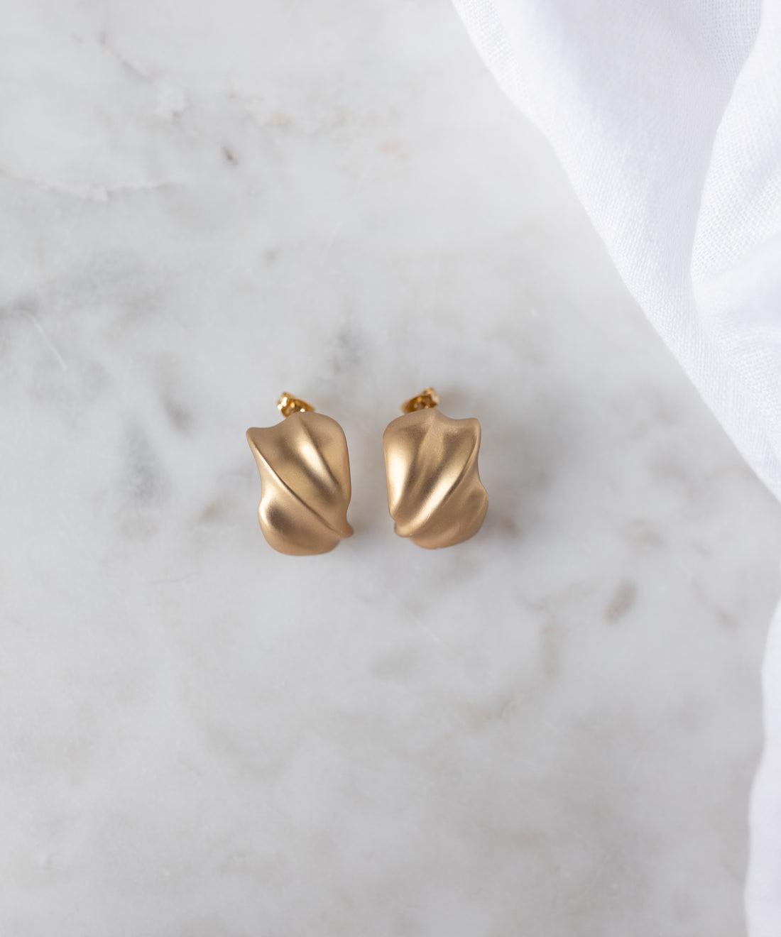 A pair of SAMPLE SALE Matt Gold Creoles by WALD Berlin, crafted in Germany with a chic abstract design, rests on white marble with fabric nearby—a stylish sample sale treasure.