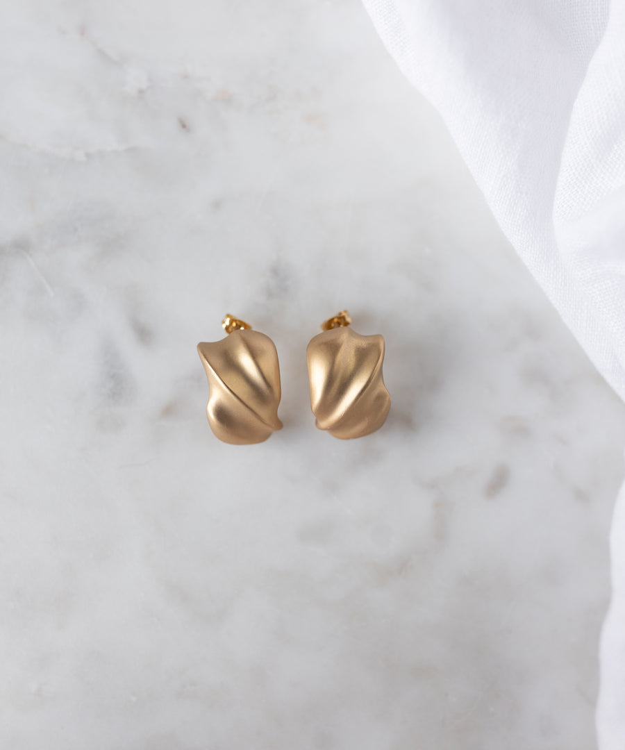 A pair of SAMPLE SALE Matt Gold Creoles by WALD Berlin, crafted in Germany with a chic abstract design, rests on white marble with fabric nearby—a stylish sample sale treasure.