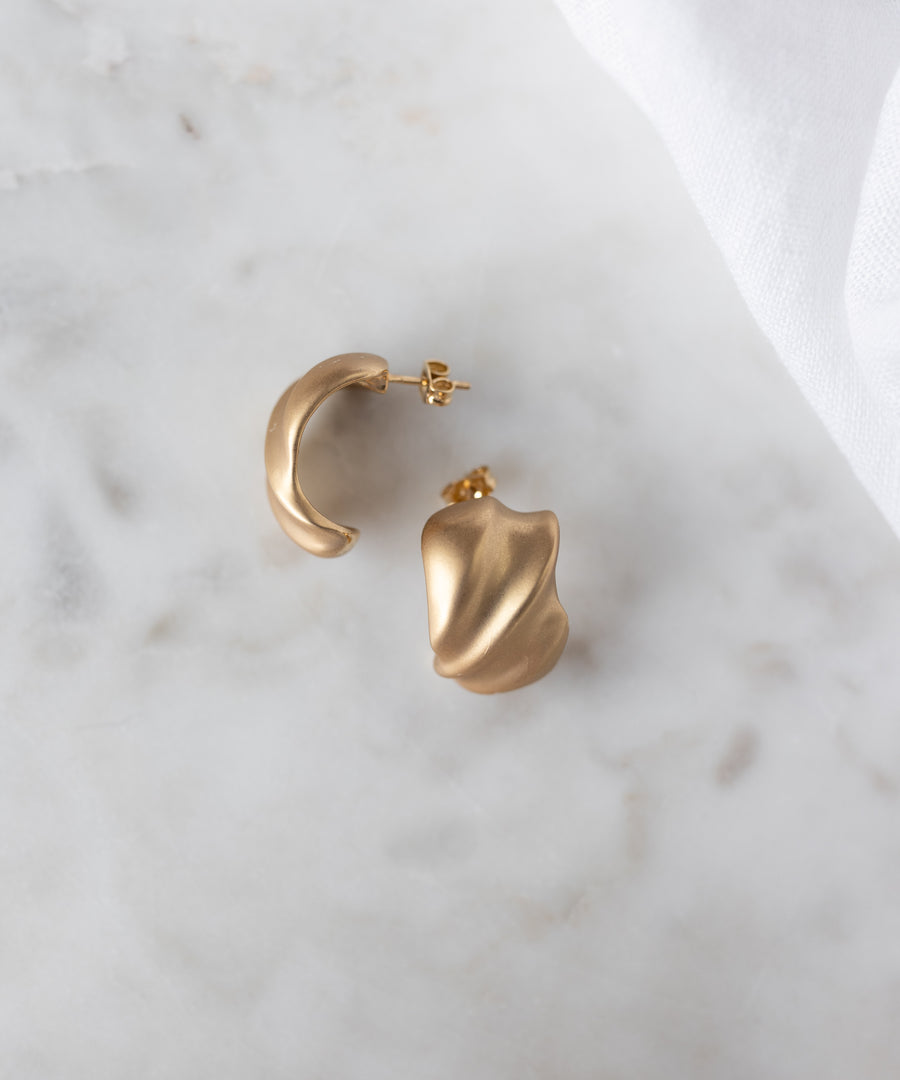 SAMPLE SALE Matt Gold Creoles by WALD Berlin—18k gold plated, abstract-shaped earrings made in Germany—displayed on white marble with a white cloth in the corner.