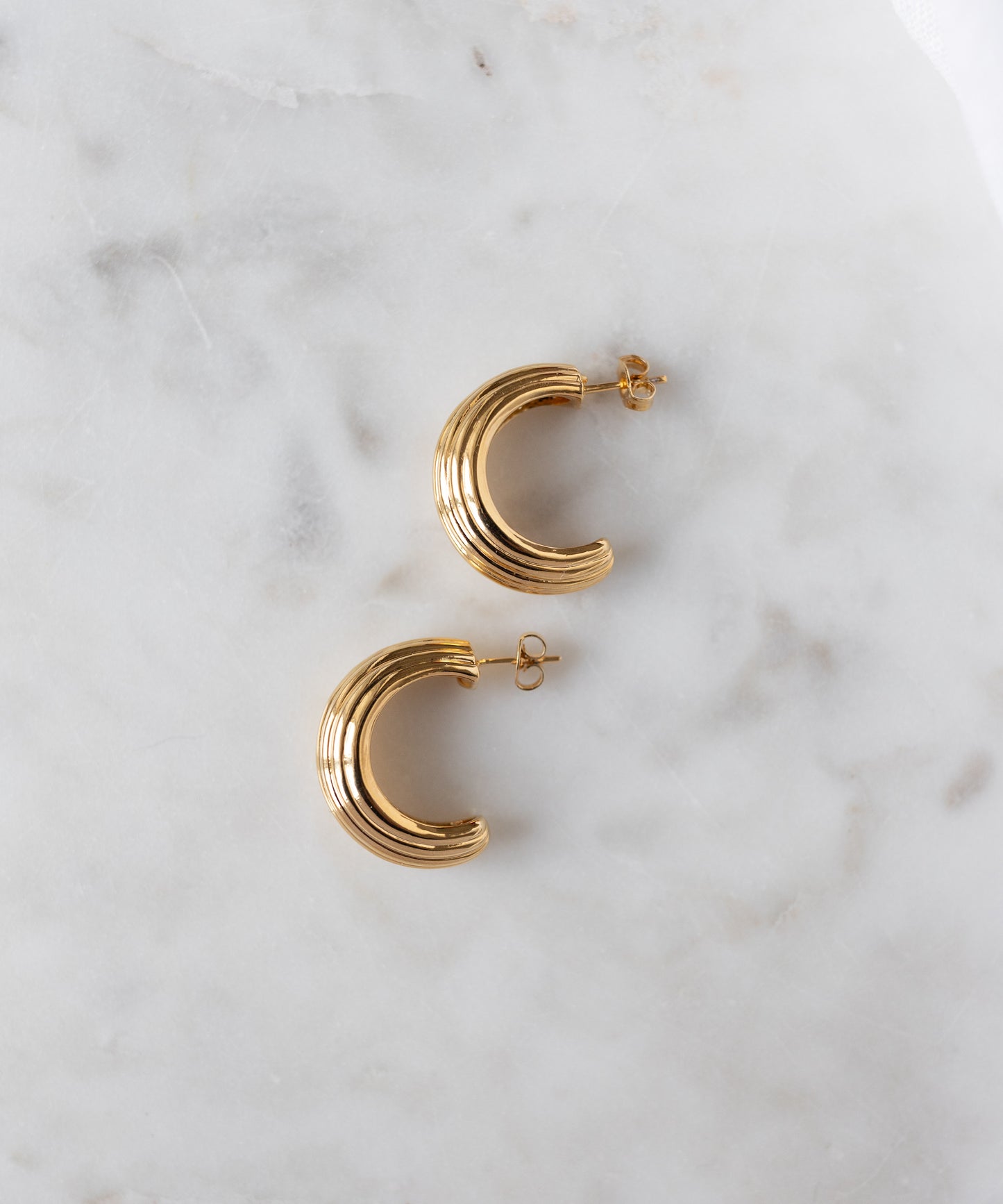 The WALD Berlin Pop Wave Earrings Big are gold plated brass creoles with a ridged design and 925 silver pin, displayed on a white marble surface.