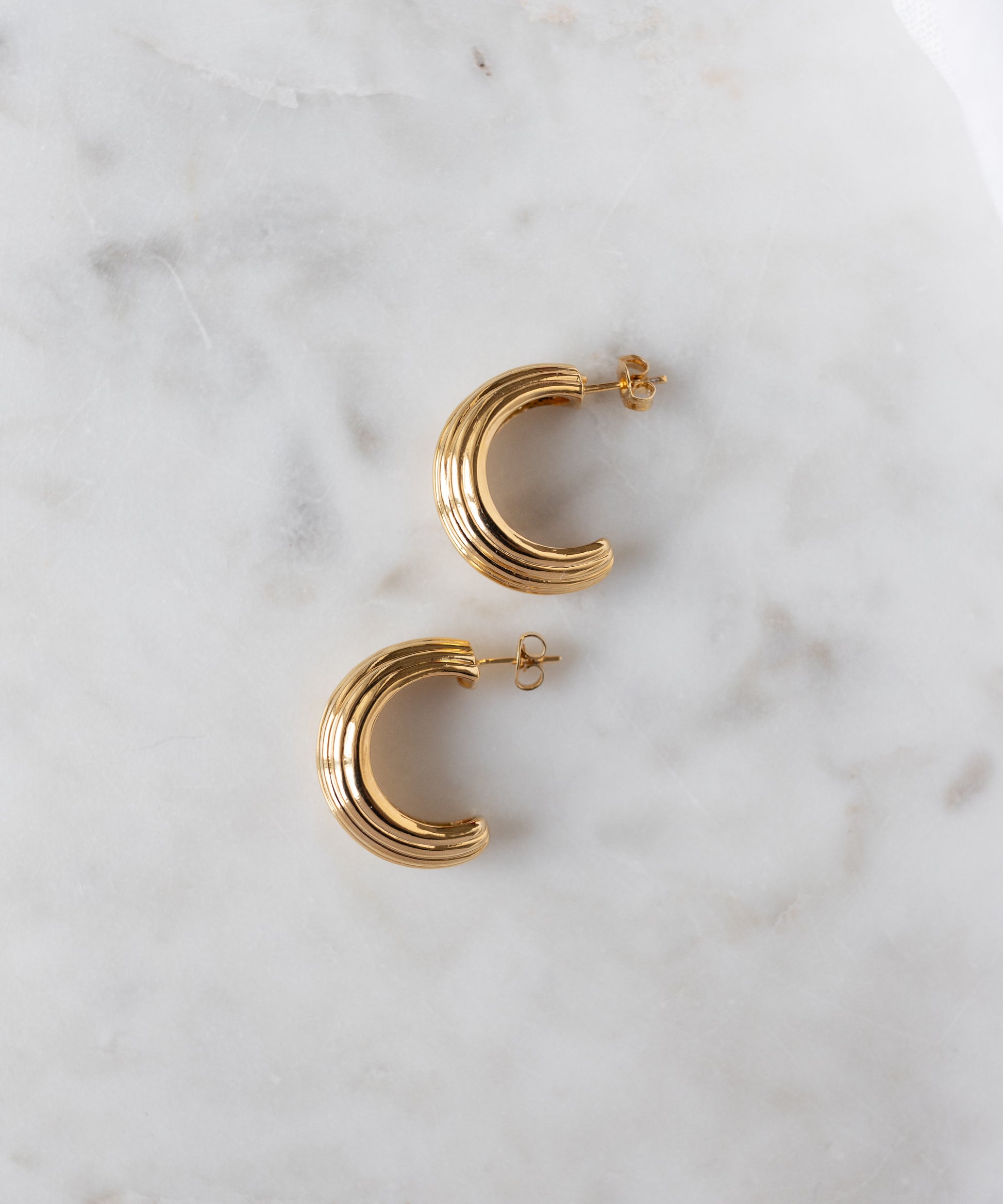 The WALD Berlin Pop Wave Earrings Big are gold plated brass creoles with a ridged design and 925 silver pin, displayed on a white marble surface.