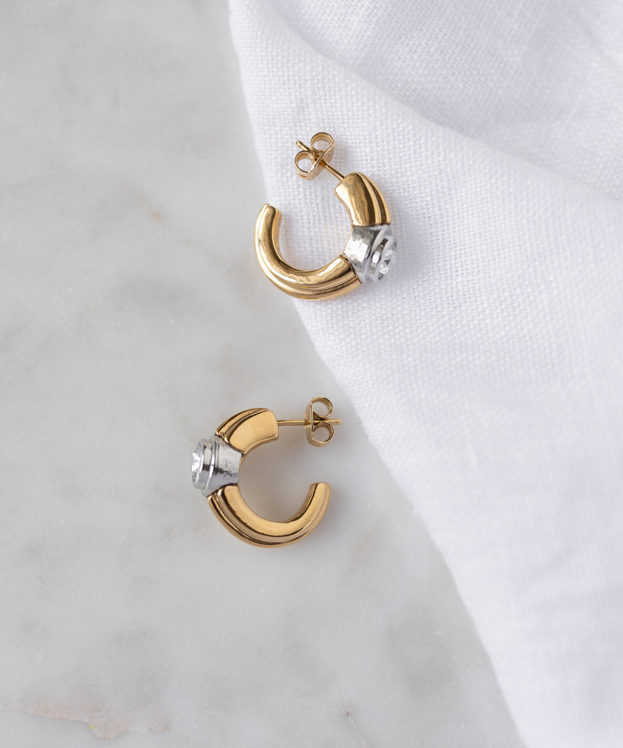 The SAMPLE SALE Pop Bi Color Crystal Earrings by WALD Berlin, with gold and silver tones, are displayed on a white marble surface partially covered by a white cloth.