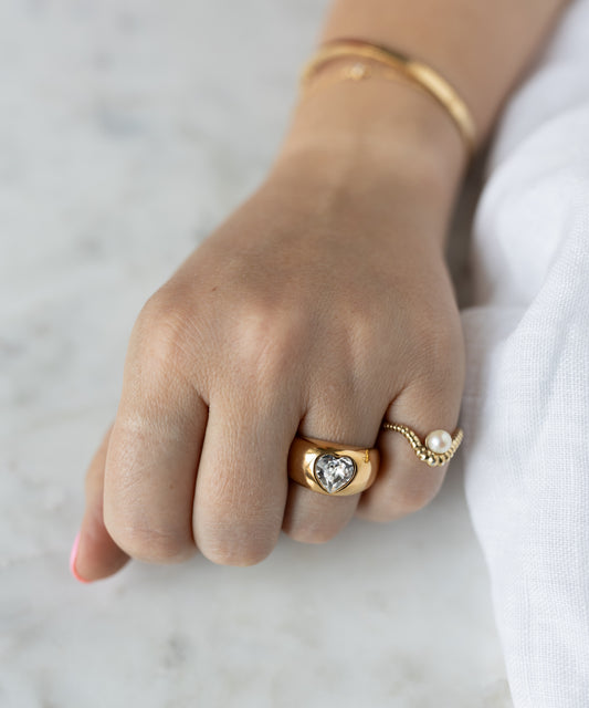 A hand wearing three gold rings, including the WALD Berlin Mom Swarovski Heart Ring and a pearl ring, plus a gold plated bracelet on the wrist, resting on a white surface.