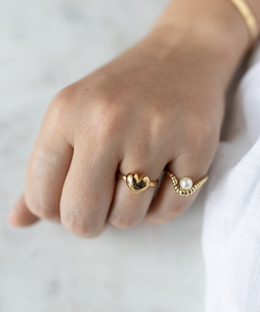 A hand wears two gold rings against a light background: the WALD Berlin Mom Heart Ring, symbolizing love, and another with a small pearl.