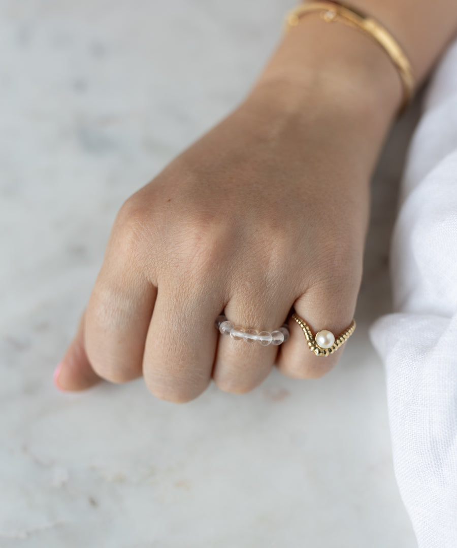 A hand wearing the WALD Berlin Pop Gemstone Ring Set—featuring clear beads, a gold ring with pearl—and a gold bracelet rests on light marble, making it perfect for gift inspiration.