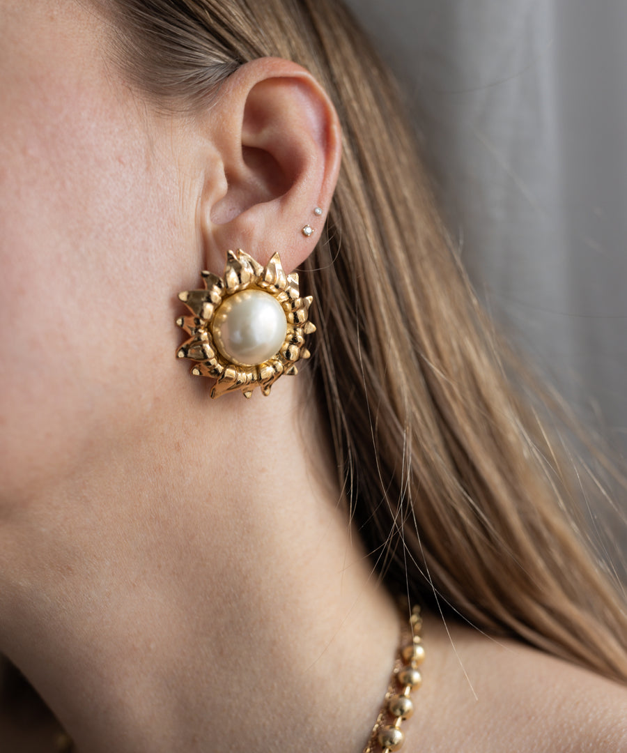 Close-up of a woman's ear wearing WALD Berlin SAMPLE SALE Pop Soleil Earrings—large, gold plated with pearl flower detail—paired with two small studs and long brown hair visible; a striking statement jewelry piece.