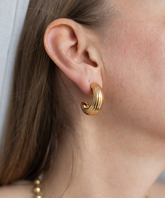 Close-up of a person's ear wearing WALD Berlin's Pop Wave Earrings Big, thick gold-plated brass spirals; brown hair and part of a gold necklace are also visible.