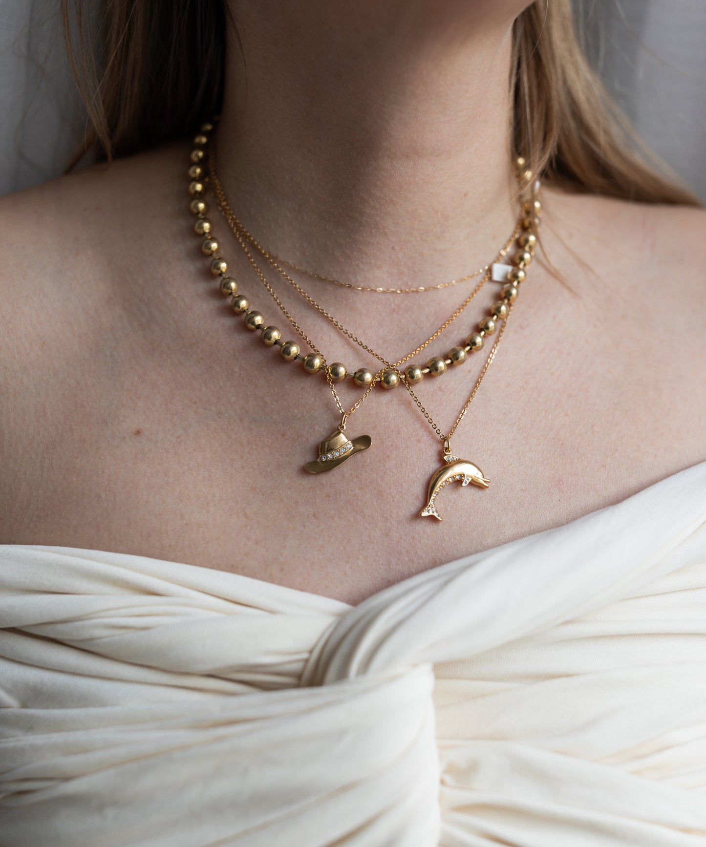 A person wears layered gold-plated brass necklaces, including the WALD Berlin Pop Dolphin Necklace, with an off-shoulder white gathered fabric top.