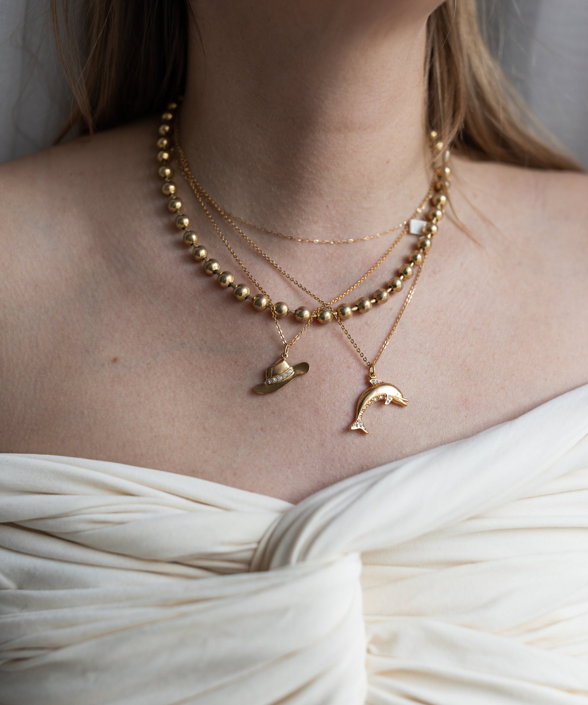 A person wears layered gold-plated brass necklaces, including the WALD Berlin Pop Dolphin Necklace, with an off-shoulder white gathered fabric top.