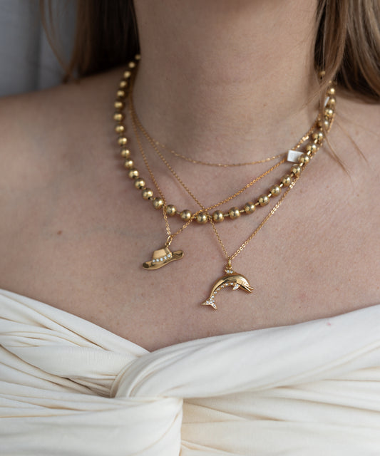 A woman in an off-white knotted top wears WALD Berlin's Pop Dolphin Necklace—layered goldplated brass with dolphin and planet pendants.