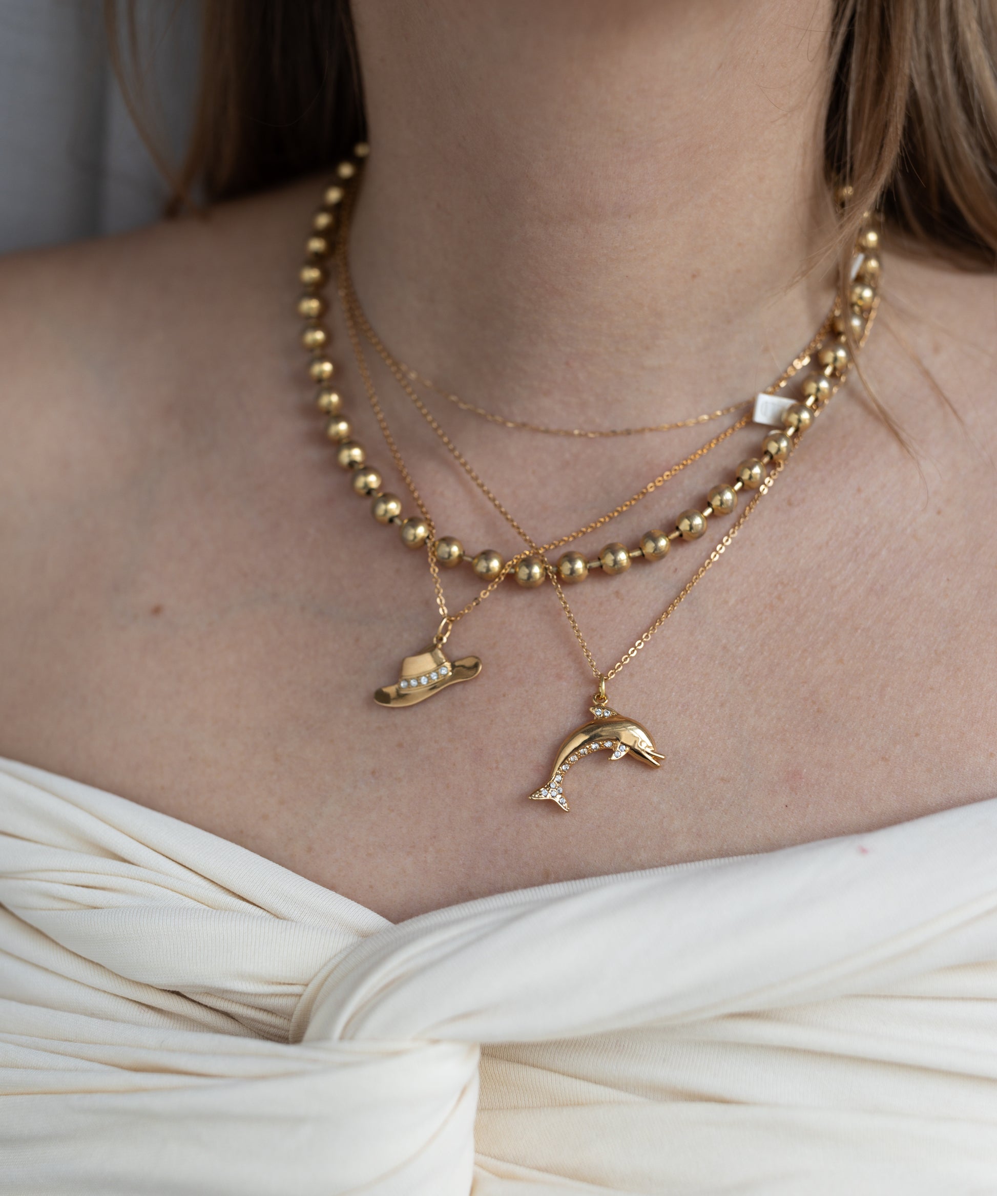 A woman in an off-white knotted top wears WALD Berlin's Pop Dolphin Necklace—layered goldplated brass with dolphin and planet pendants.