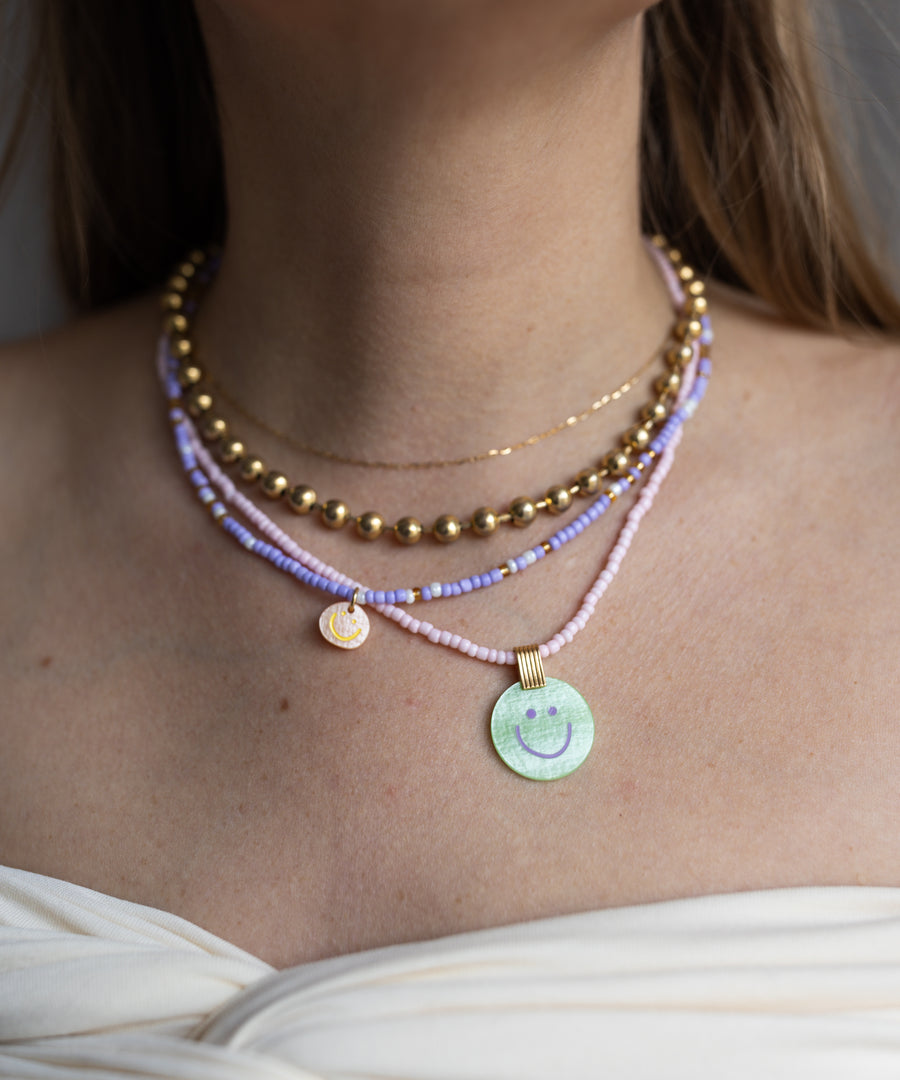 A woman layers WALD Berlin necklaces, featuring pastel beads and 18k gold-plated strands with two Pop Smilie Medallion Green pendants, over a white off-shoulder top for a fresh summer look.