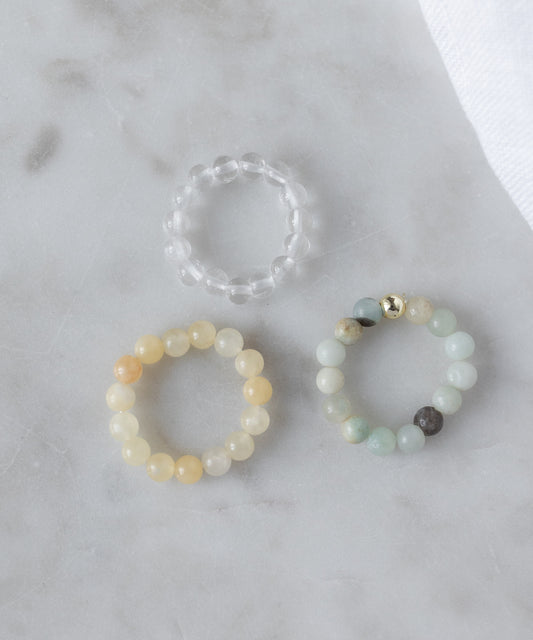 The WALD Berlin Pop Gemstone Ring Set features three beaded rings—a clear gemstone, a yellow band, and a multicolored ring with a gold bead—arranged on light marble. Their flexible sizing makes them charming gift ideas.