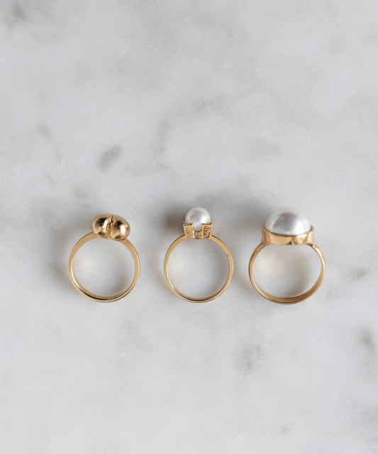 The WALD Berlin Mom Heart Ring, crafted from gold plated brass and featuring unique pearl or bead settings, is displayed on light marble with two other gold rings, radiating elegance and love.