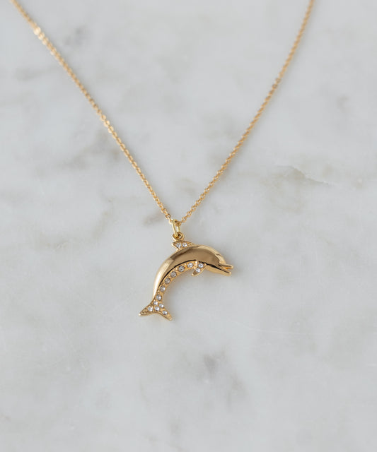 The WALD Berlin Pop Dolphin Necklace, a goldplated brass piece with a dolphin-shaped pendant embellished with sparkling Swarovski stones, is elegantly displayed on a light marble surface.