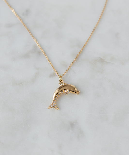 The WALD Berlin Pop Dolphin Necklace, a goldplated brass piece with a dolphin-shaped pendant embellished with sparkling Swarovski stones, is elegantly displayed on a light marble surface.