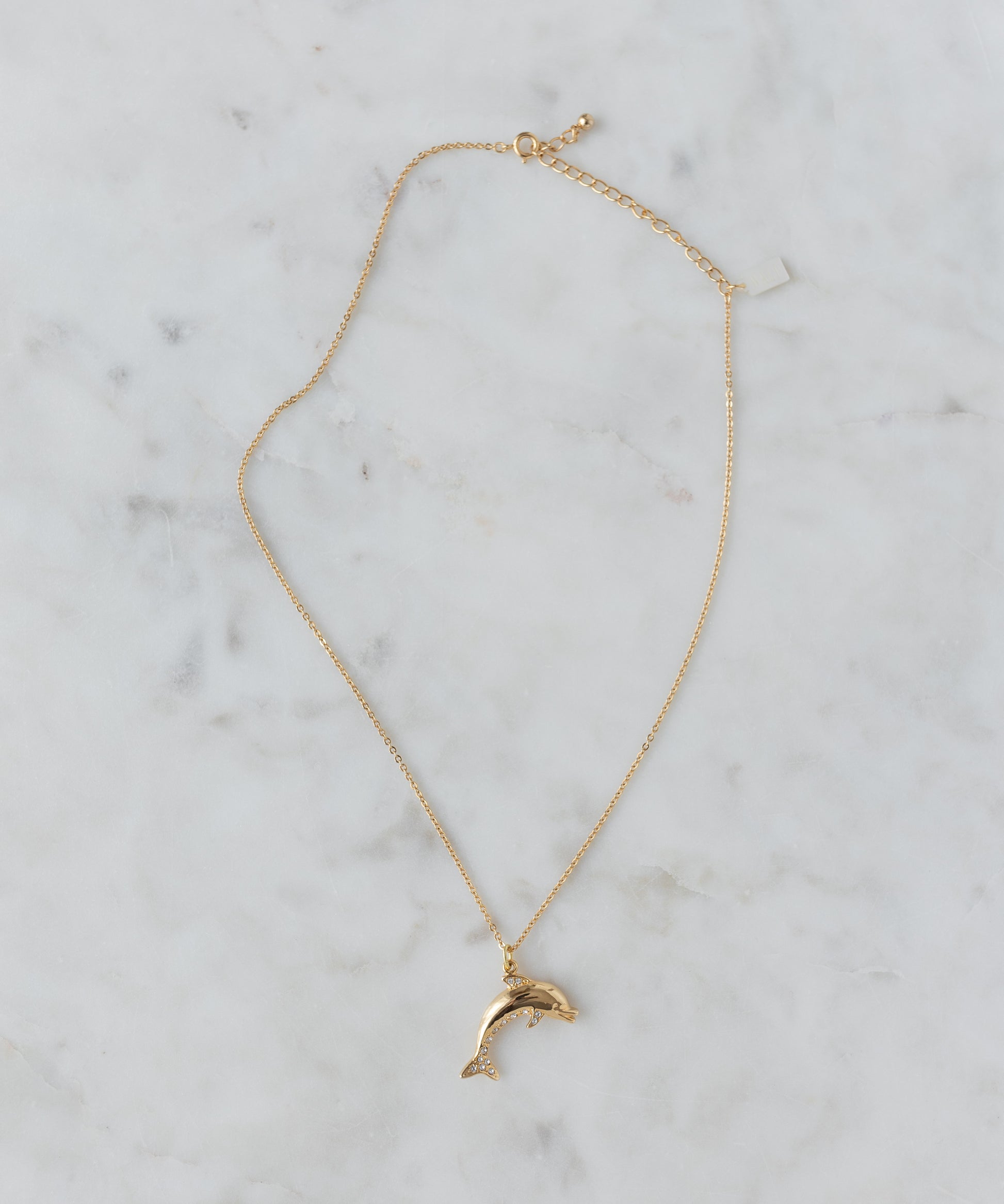 The WALD Berlin Pop Dolphin Necklace features a dolphin-shaped pendant in gold-plated brass, adorned with sparkling Swarovski stones, and is shown on a light marble surface.