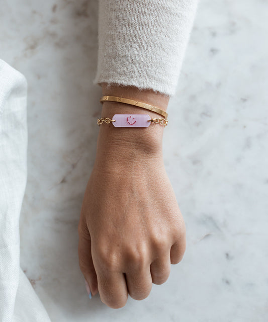 A hand wears two 18k gold plated bracelets and WALD Berlin's Happy ID Rose Bracelet exclusive—a delicate chain with a pink tag and simple smiley face—resting on marble.
