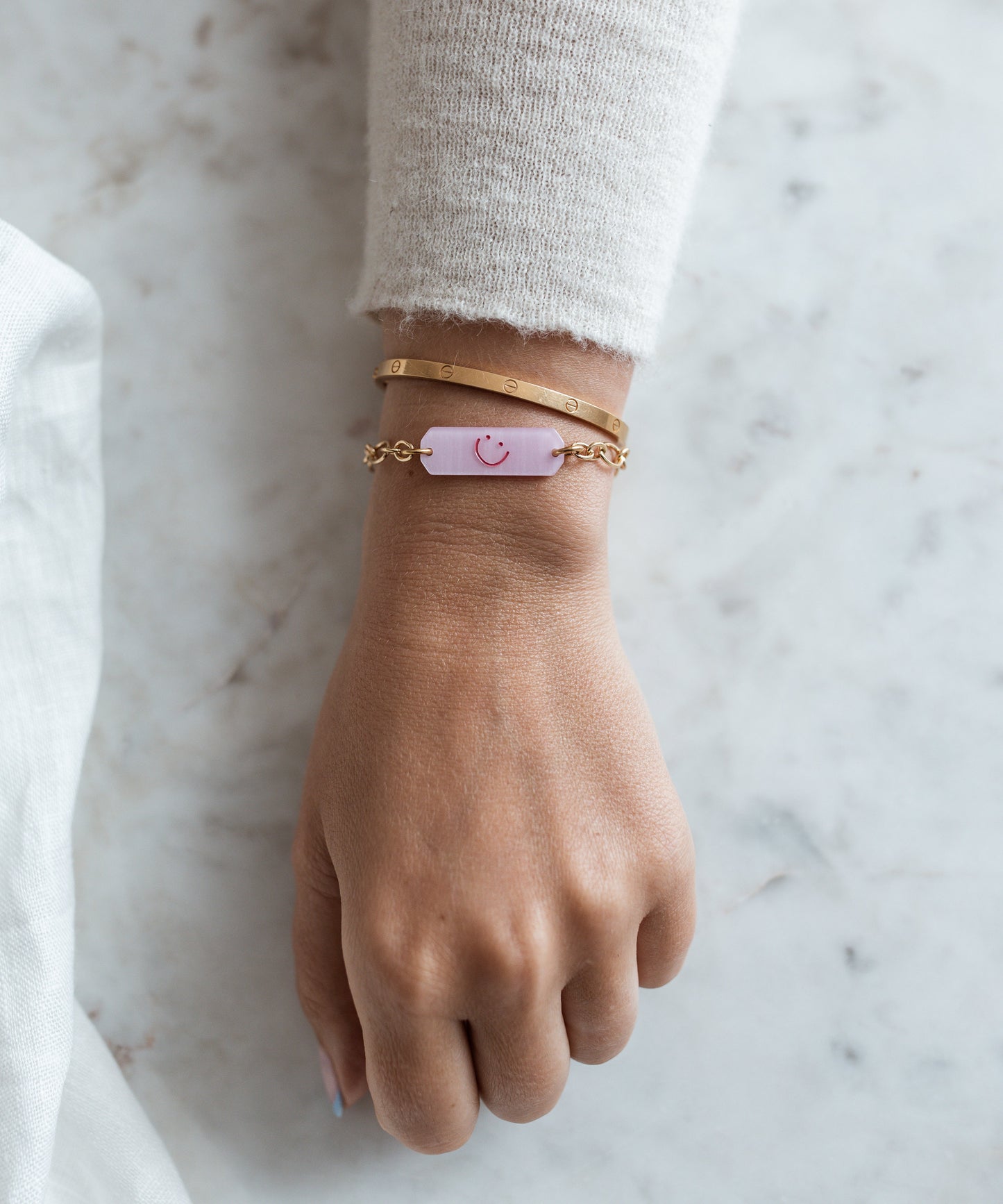 A hand wears two 18k gold plated bracelets and WALD Berlin's Happy ID Rose Bracelet exclusive—a delicate chain with a pink tag and simple smiley face—resting on marble.