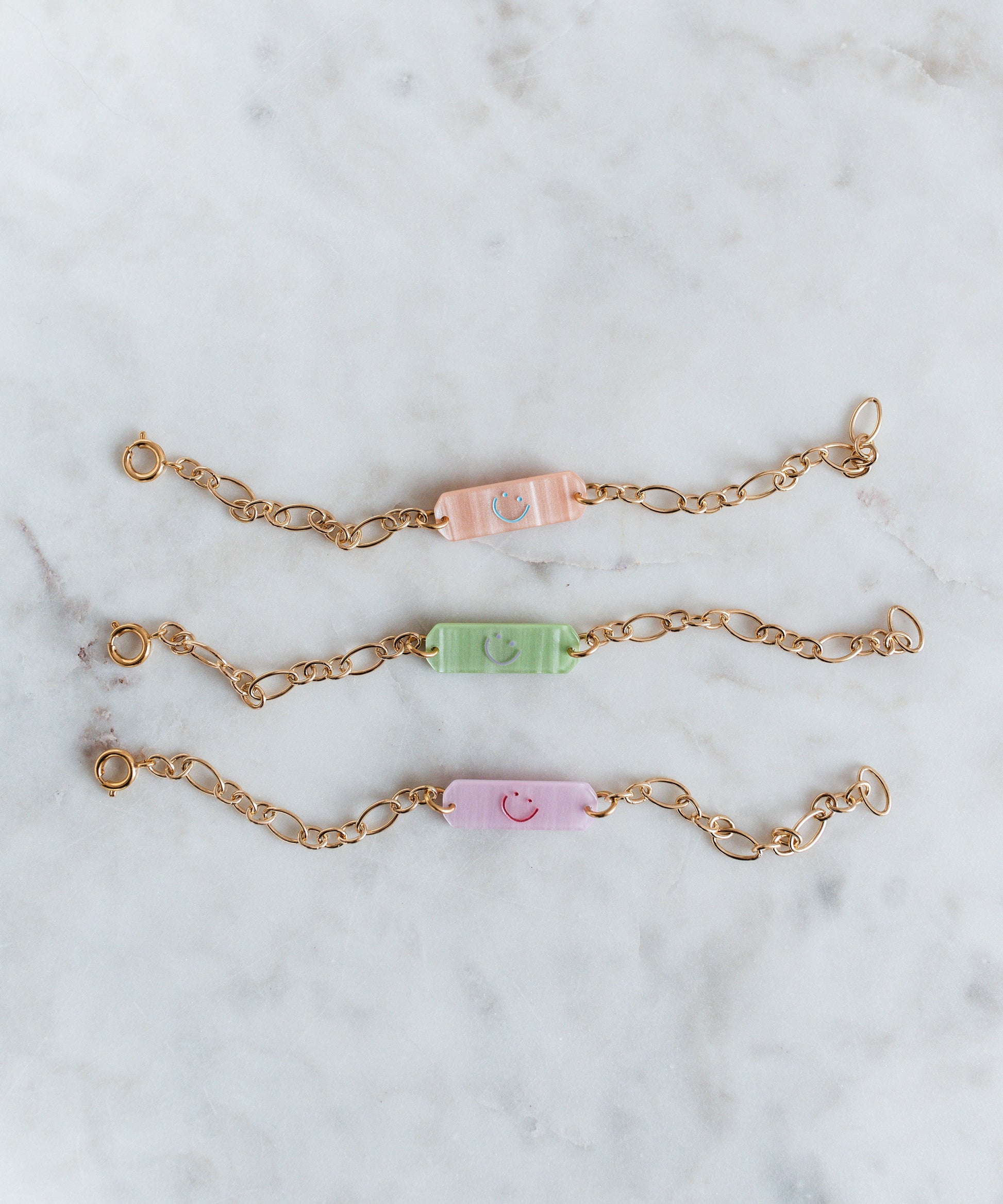 The Happy ID Rose Bracelet exclusive by WALD Berlin features three 18k gold plated chain bracelets with pastel rectangular beads in peach, green, and lavender, each accented with a smiley face, displayed on white marble.
