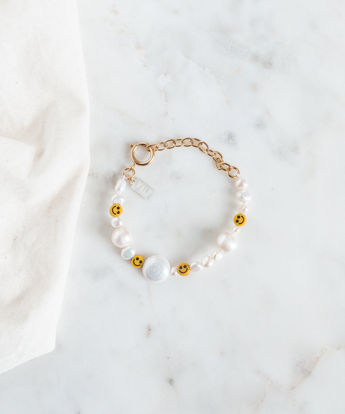 The WALD Berlin Smilie Dude Pearl Bracelet with Extention is showcased on a marble surface, featuring sweetwater pearls and cheerful smiley beads.