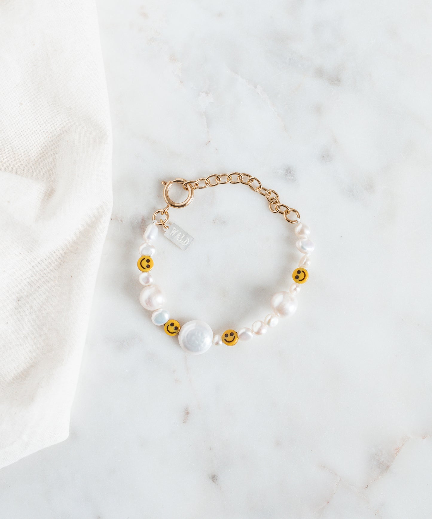 The WALD Berlin Smilie Dude Pearl Bracelet with Extention is showcased on a marble surface, featuring sweetwater pearls and cheerful smiley beads.