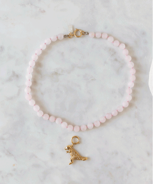 Eine rosa Perlenkette mit einem goldenen Dinosaurier-Anhänger aus dem „DIY-Schmuck-Workshop x MINI im KaDeWe Berlin“ von WALD Berlin wird auf einer hellen Marmorfläche präsentiert – ideal für alle, die gerne Schmuck basteln.